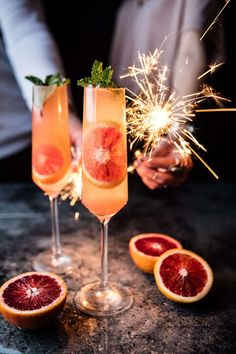 two wine glasses filled with liquid and garnished with sparklers next to grapefruits