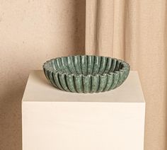 a green bowl sitting on top of a white pedestal in front of a beige curtain