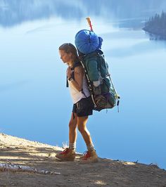 a woman with a large backpack is walking on the edge of a cliff overlooking a body of water