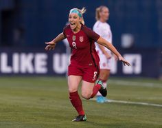 a women's soccer player is running on the field with her arms out and mouth wide open