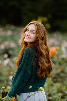 a woman with long red hair is standing in a field full of flowers and smiling at the camera