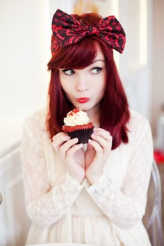 a woman with red hair holding a cupcake in front of her face and wearing a bow