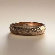 a gold wedding band with leaves engraved on the side and initials in the middle, sitting on a white surface