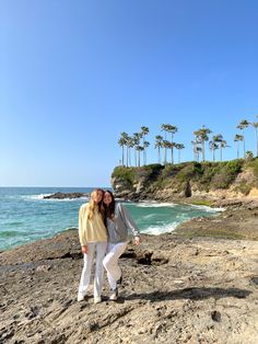 two women standing on rocks near the ocean with palm trees in the backgroud