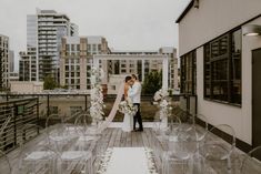 a bride and groom are standing on a rooftop with clear acrylic chairs in front of them