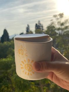 a person holding up a coffee cup in front of the sun and trees on a sunny day