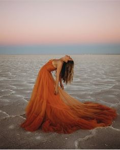 a woman in an orange dress is standing on the beach at sunset with her head down