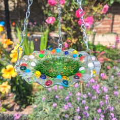 a glass bird feeder hanging from a chain in front of some flowers and other plants