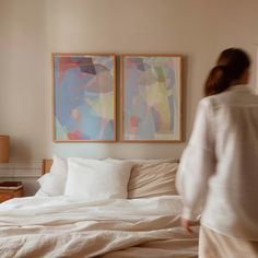 a woman is walking in front of two paintings on the wall above a white bed