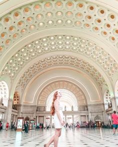 a woman in white dress walking through a large building with arches and ceiling lights above her