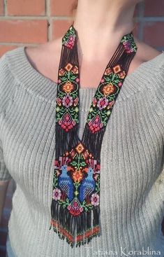 a woman wearing a gray sweater and black necklace with colorful flowers on the front, standing in front of a brick wall