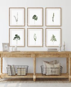 a wooden table topped with baskets under four framed plants on white wall above it is a shelf filled with books and other items