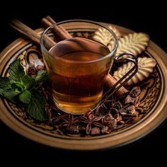 a glass cup filled with tea next to cinnamons and green leaves on a plate