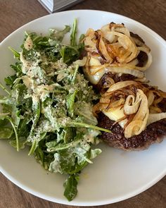 a white plate topped with meat covered in onions next to a green leafy salad