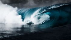 a large wave crashing into the ocean on a cloudy day with dark clouds in the background