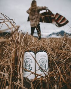 two cans of beer sitting in the middle of tall grass with a woman standing behind them