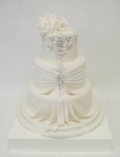 a three tiered wedding cake with white flowers and ribbons on the top, sitting on a pedestal