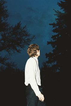 a young man standing in front of trees at night with his head turned to the side