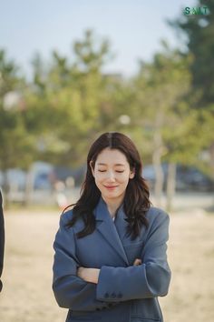 a woman with her arms crossed standing in front of some trees and sand, wearing a blue suit