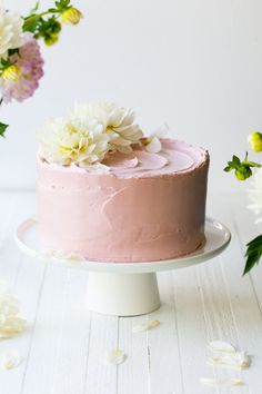 a pink frosted cake with white flowers on top sitting on a plate next to some daisies