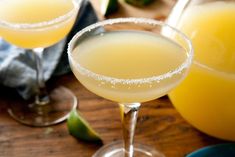 two margaritas sitting on top of a wooden table next to glasses with drinks in them