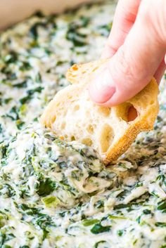 a hand dipping a piece of bread into a bowl of spinach and cheese dip