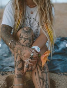 a woman sitting in the sand with tattoos on her arms and legs, holding an orange flower