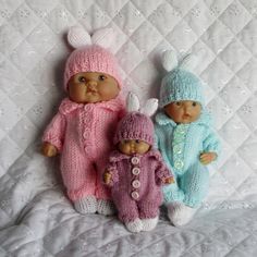 three baby dolls are laying on a white quilted bed, wearing knitted outfits