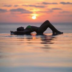 a woman laying on top of a body of water at the beach in front of a setting sun