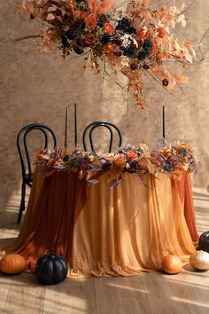 an arrangement of flowers and candles on a table with orange drapes, pumpkins and black chairs