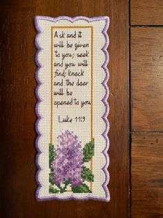 a cross stitch bookmark with a purple flower and verse on the front, sitting on a wooden surface