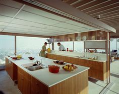 two people standing in a kitchen next to a sink and counter top with fruit on it