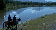 two people riding horses near a lake and mountains in the distance with trees on either side