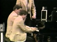 two men are playing the piano while another man stands behind them in front of him