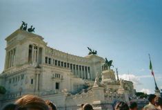 a crowd of people standing in front of a large building with statues on the top