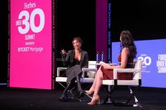 two women sitting on chairs talking to each other in front of a pink screen with the number 30