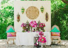 a table with flowers and lanterns on it in the middle of a stone patio surrounded by greenery