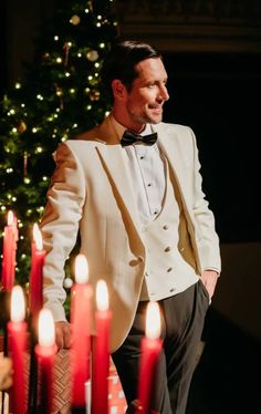 a man in a tuxedo standing next to candles with a christmas tree behind him