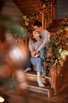 a man and woman sitting on the steps in front of christmas lights with their arms around each other