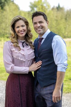 a man and woman standing next to each other in front of a green field with trees