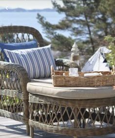 a wicker chair with pillows and a basket on it sitting on a patio next to the ocean