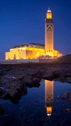 the building is lit up at night with its reflection in the water and on the rocks