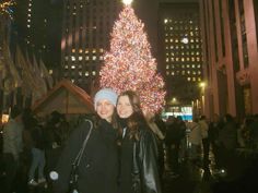 two women standing next to each other in front of a large christmas tree at night