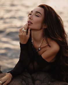 a beautiful woman sitting on top of a rock next to the ocean with her hand in her mouth