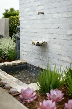 a water fountain in front of a white brick wall with pink flowers and greenery