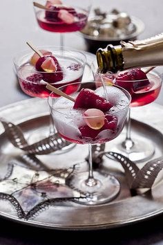 two glasses filled with red wine and cherries on a silver tray next to another glass