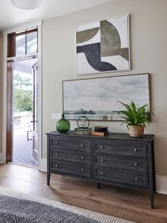 a living room with a large painting on the wall next to a black dresser and potted plant