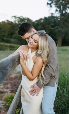 a man and woman embracing each other in front of a fence with grass behind them