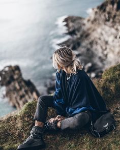 a woman sitting on top of a grass covered hillside next to the ocean with her hand in her pocket