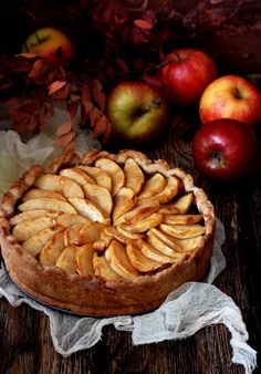 an apple pie on a wooden table with apples in the background and leaves around it